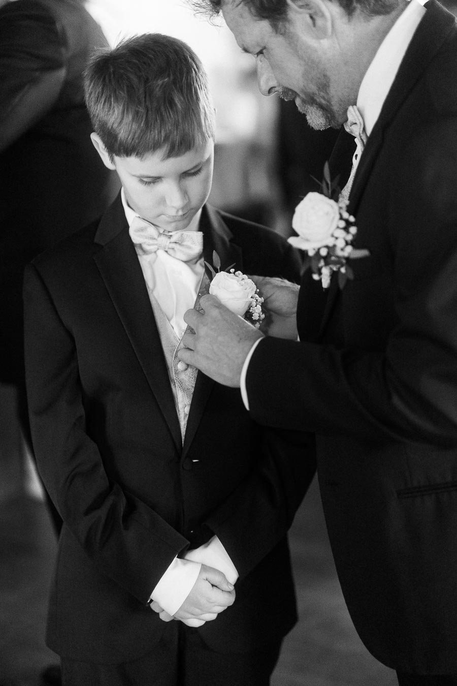 A young boy in a formal suit looks down as an older man adjusts his boutonniere, captured in a black and white photograph, highlighting a touching moment of care at a formal event.