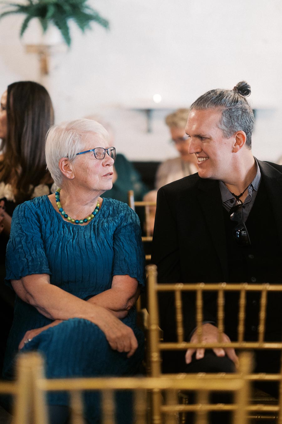 An elderly woman in blue and a man in a dark suit smiling and chatting while seated at an indoor event, surrounded by other attendees.
