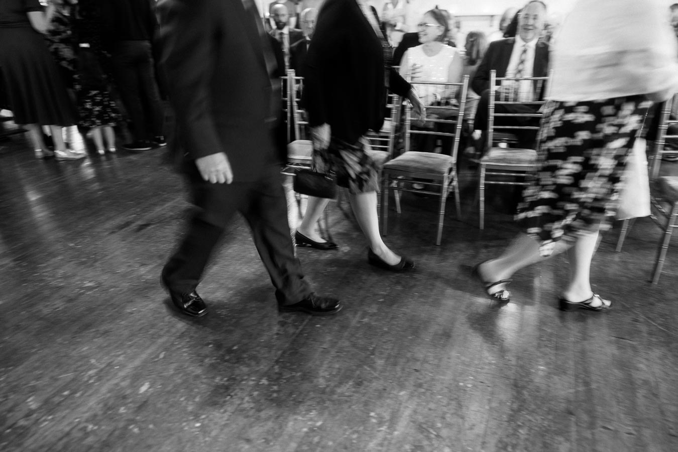 Black and white image of people walking past rows of chairs at a formal event, with attendees seated in the background.