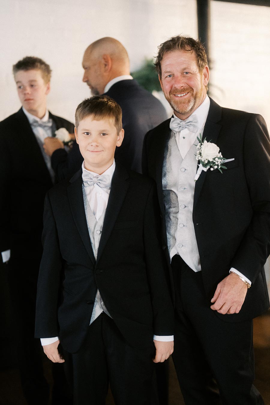 A group of men and boys dressed in formal black suits and silver waistcoats, smiling at a wedding ceremony.