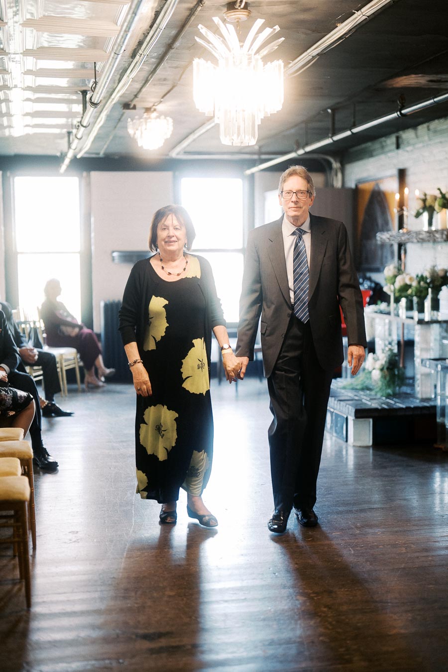 Elderly couple holding hands walking down an aisle at a formal event, with elegant chandeliers and floral decor in a warmly lit venue.