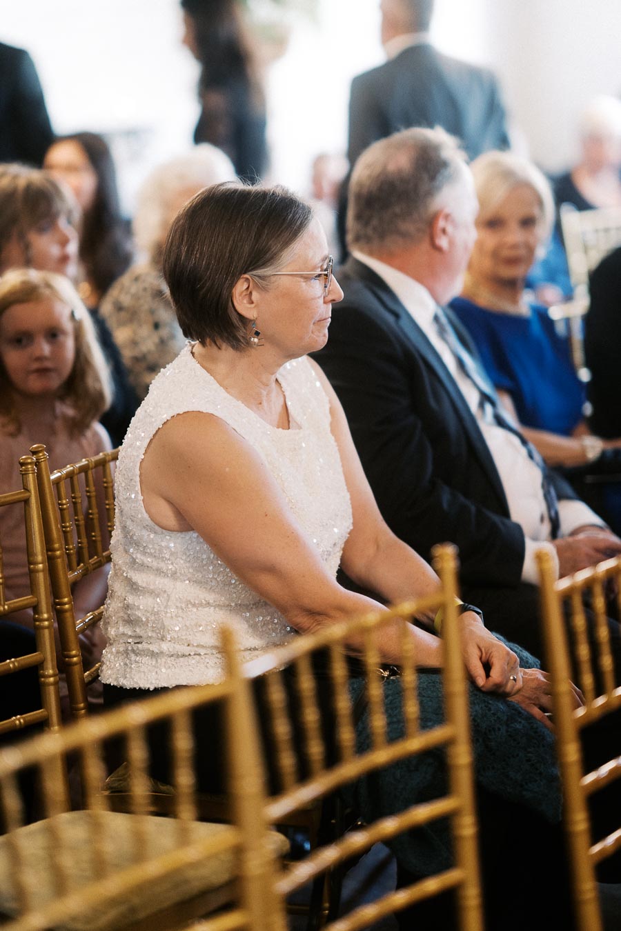 Elderly woman in a white sleeveless top seated on a gold chair at an indoor event, surrounded by other attendees.