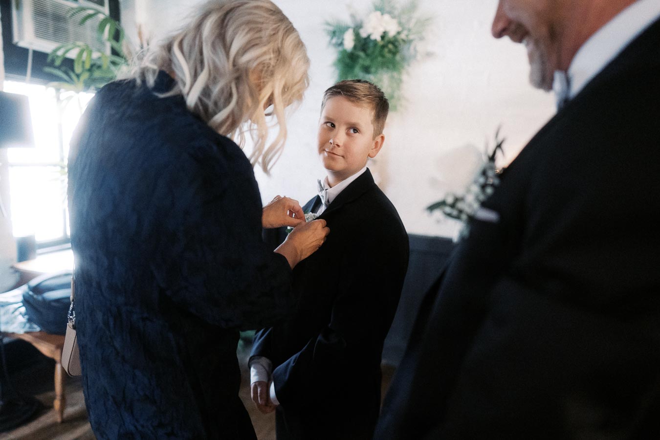 A young boy in a suit stands as an older woman adjusts his boutonniere.