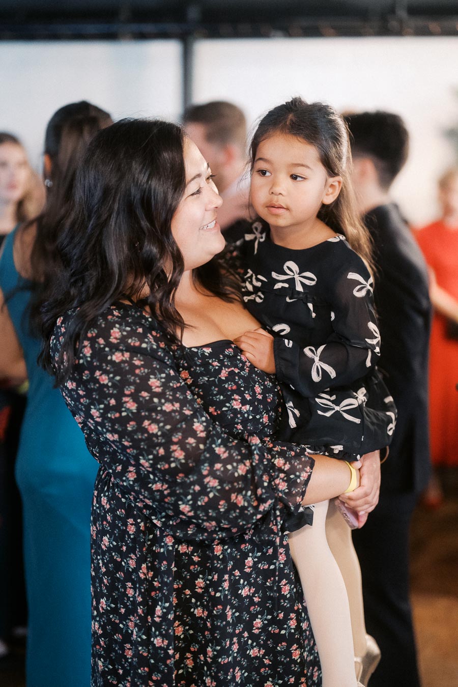 A woman in a floral dress holds a young girl wearing a dress with bow patterns at a social gathering, surrounded by other people.