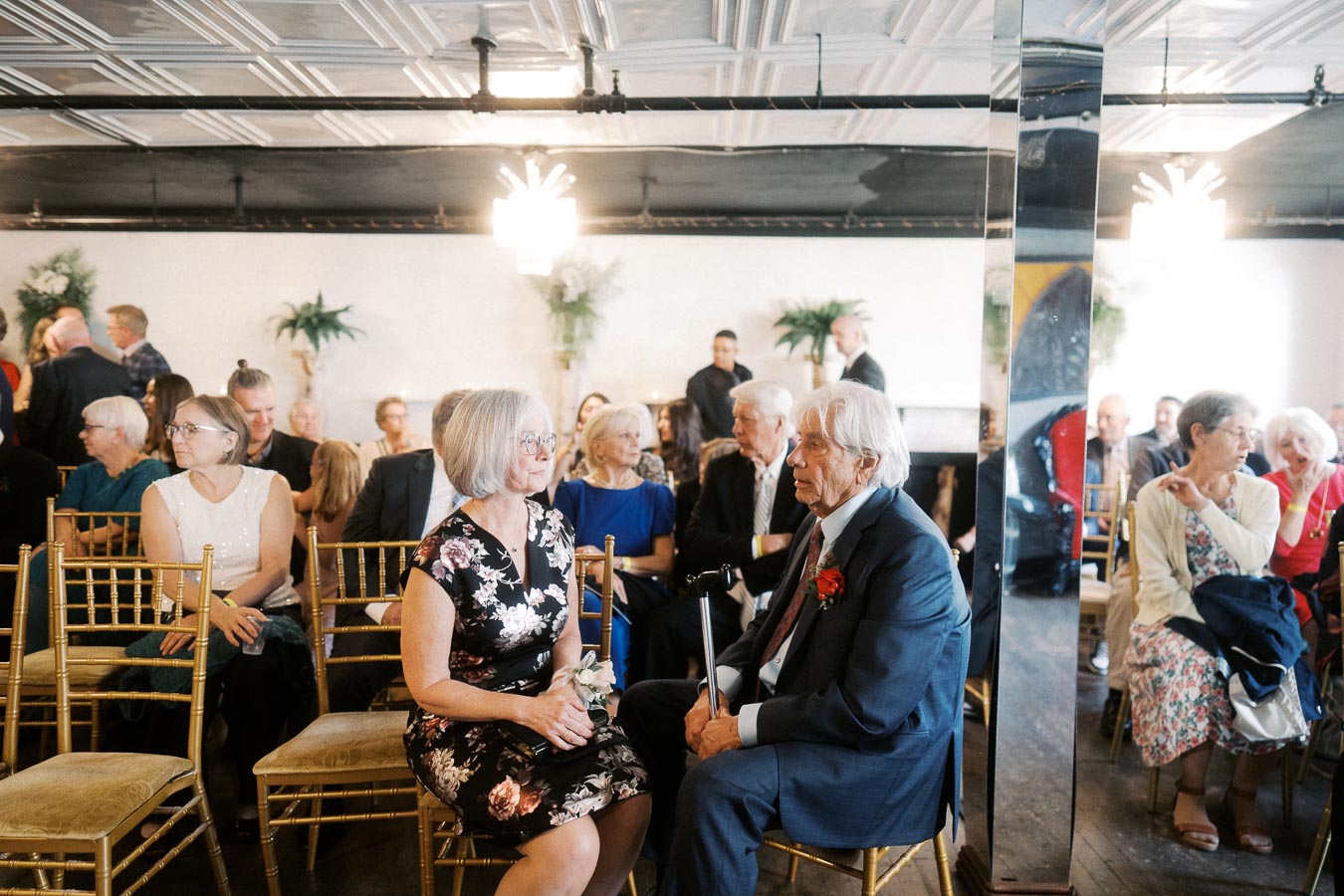 Elderly couple in formal attire conversing at a social event with guests seated in rows in a decorated hall.