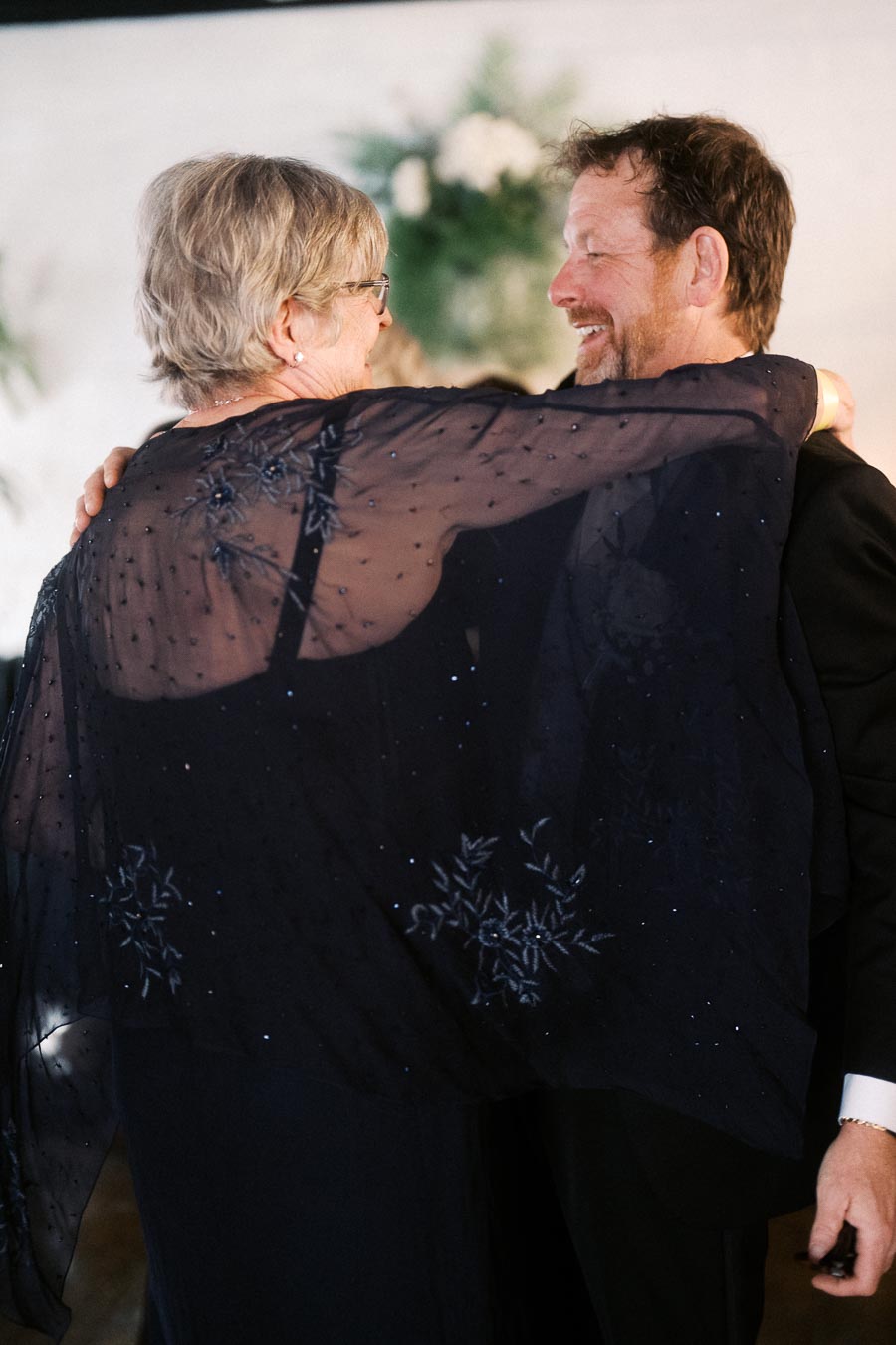 Elderly couple smiling and dancing together at a festive event, showcasing joy and togetherness, with a floral backdrop.