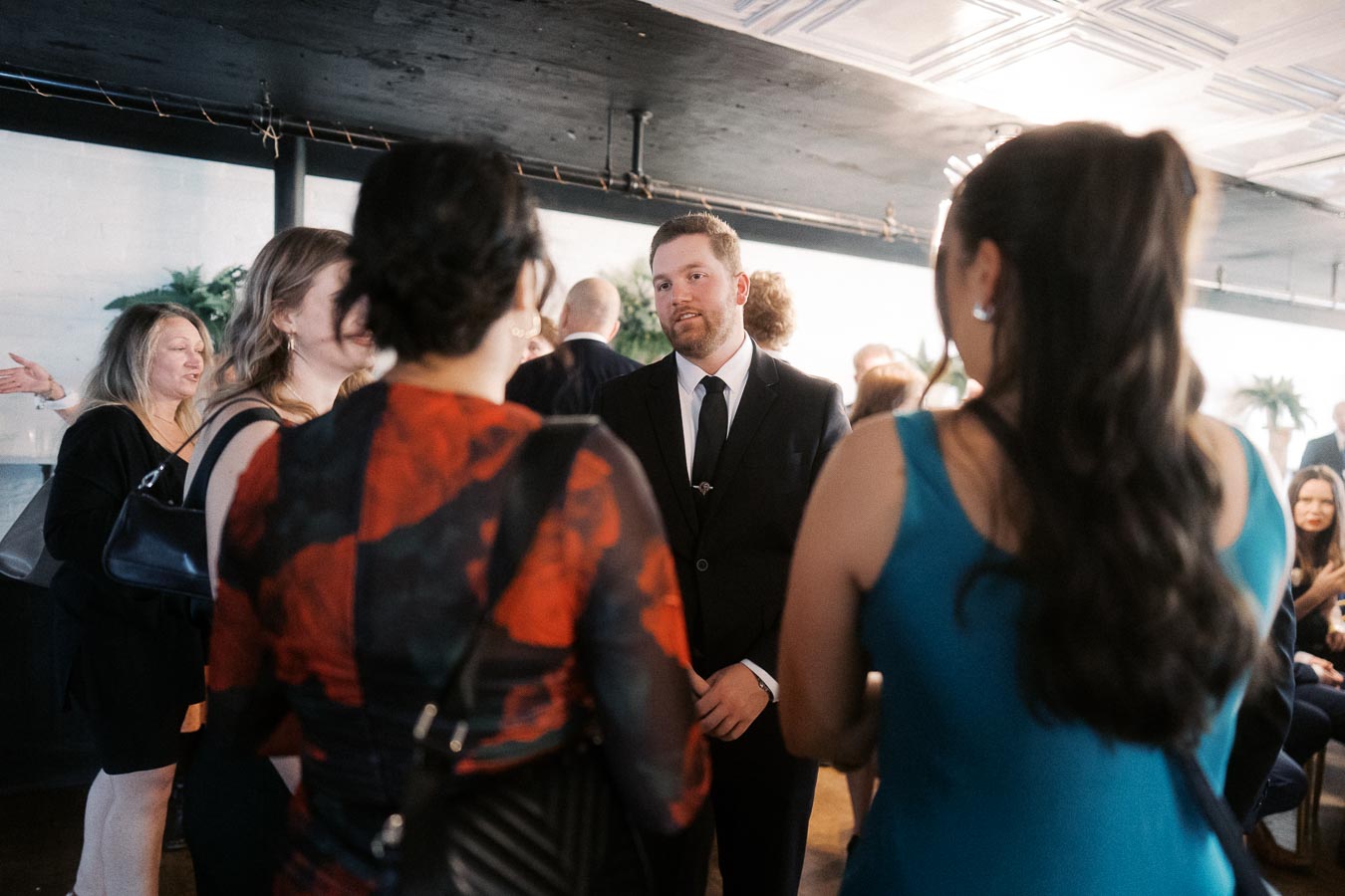 Group of people socializing at a formal event in a stylish indoor setting, with a man in a suit conversing with two women in vibrant dresses.