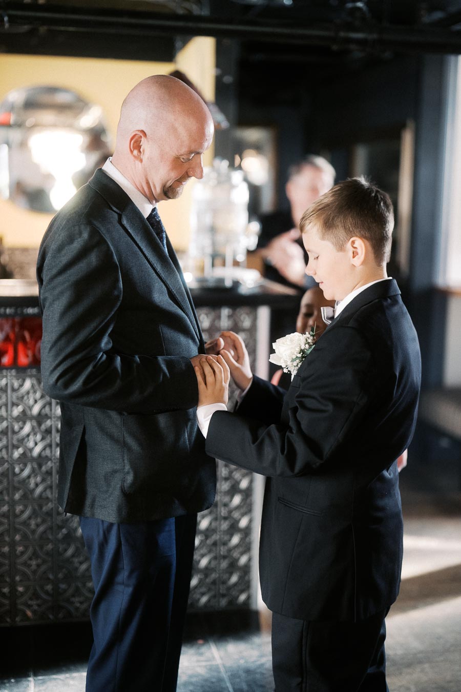 A man and a young boy wearing formal suits, sharing a heartfelt moment in a warmly lit room. The boy, with a white floral boutonniere, appears attentive as they interact in a bonding gesture.