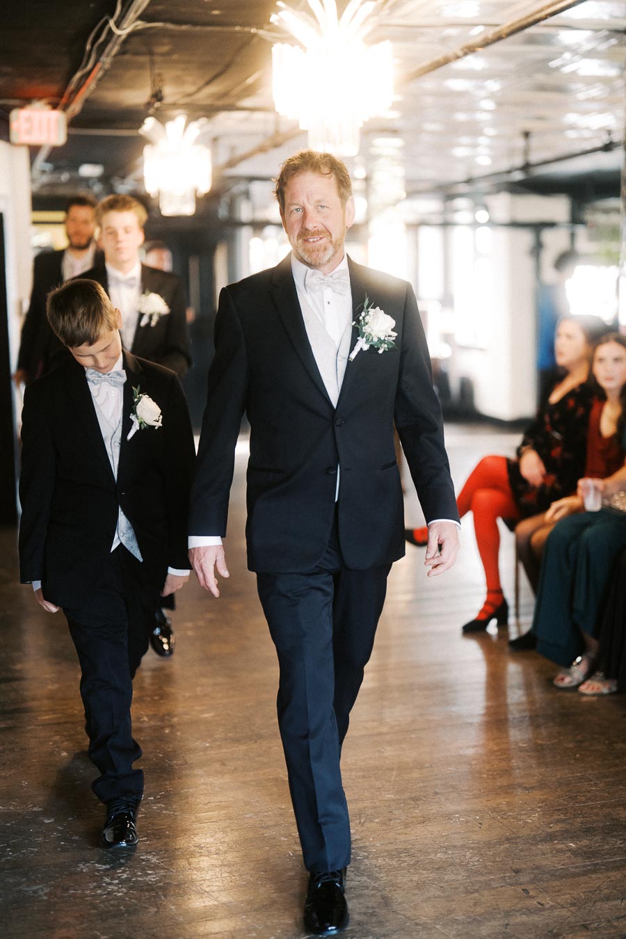 A group of men in formal suits with boutonnières walking down an aisle, captured in a well-lit indoor setting with chandeliers, during a wedding ceremony.