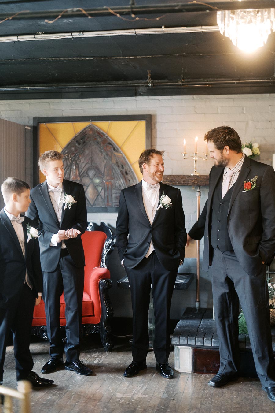 A group of four men, including a groom and groomsmen, standing indoors in formal black suits with white boutonnieres, smiling and engaging in conversation. The setting features elegant decor, including a red chair and a candle stand, contributing to a sophisticated atmosphere.
