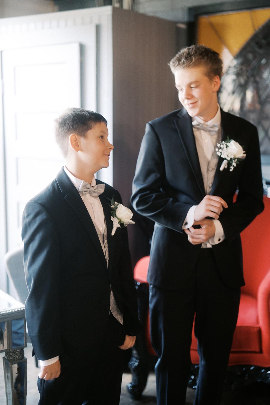 Two young boys dressed in formal black suits with bow ties and white boutonnieres, standing and smiling at each other in a well-lit indoor setting, highlighting their elegant attire for a formal event or occasion.
