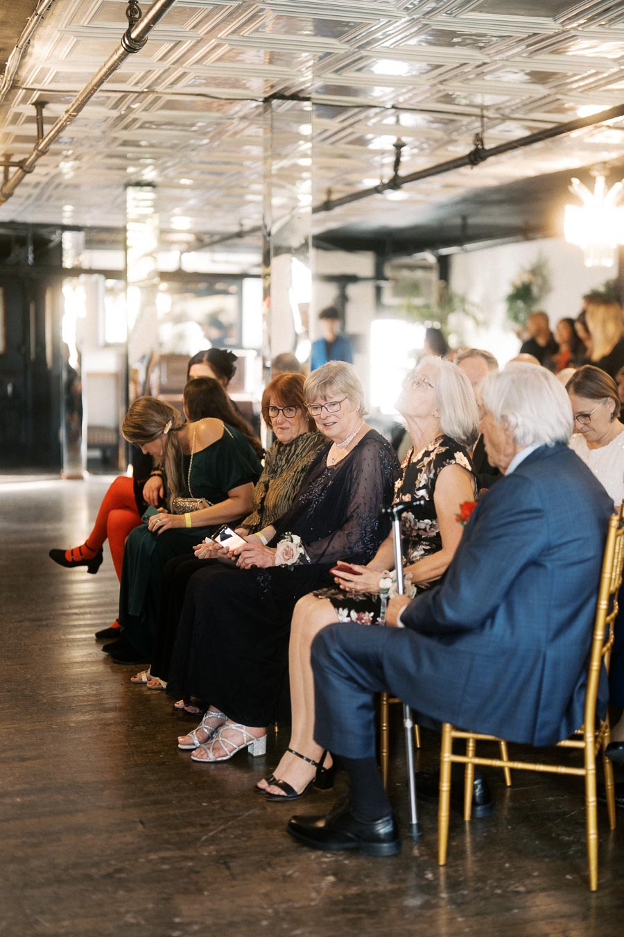 Guests seated at an indoor event, dressed in formal attire, engaging with each other in a warmly lit venue with elegant decor and reflective ceiling.