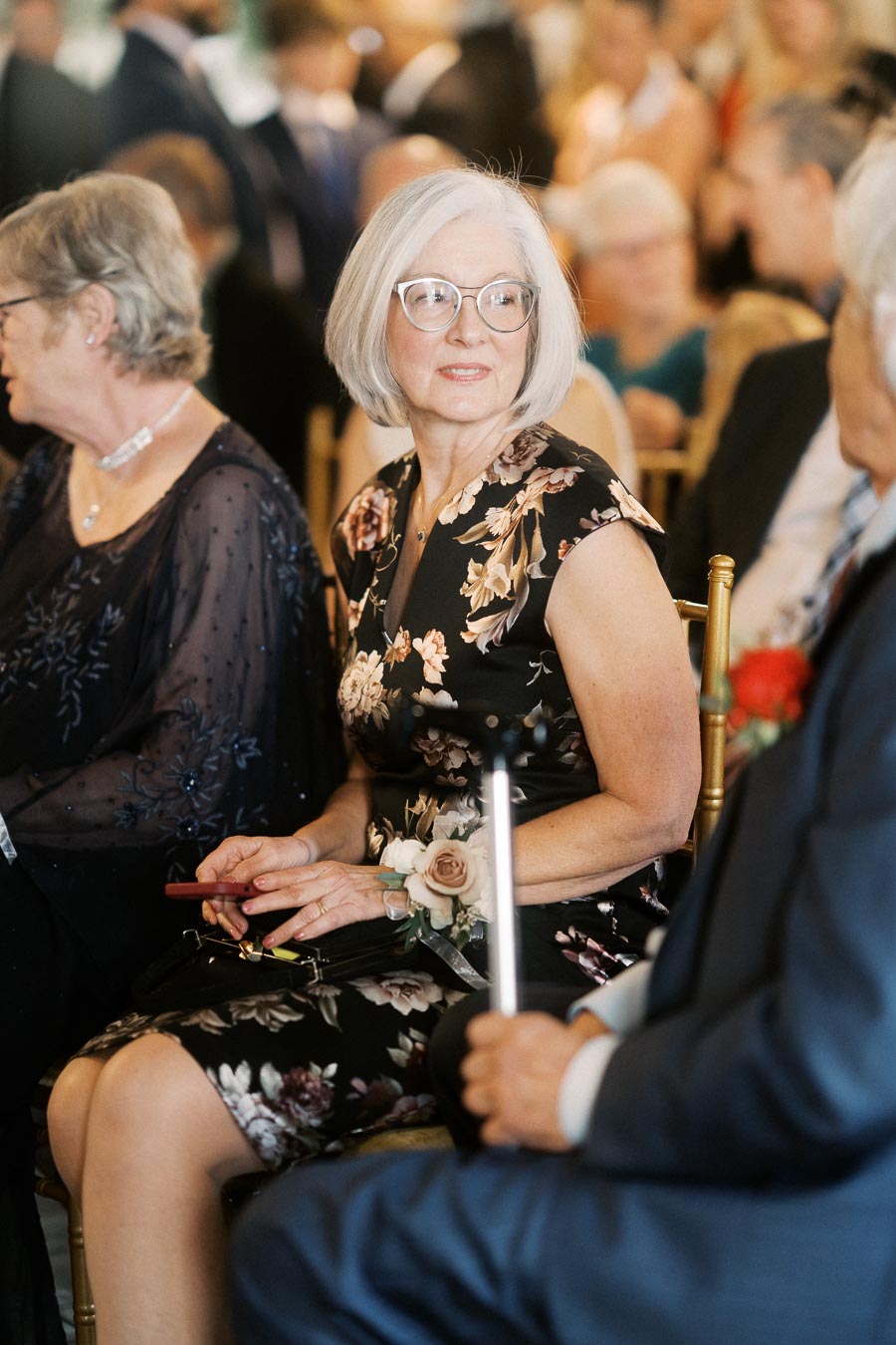 Elderly woman with short gray hair wearing glasses and a floral dress, sitting among a crowd at an indoor event, holding a red phone and floral corsage.