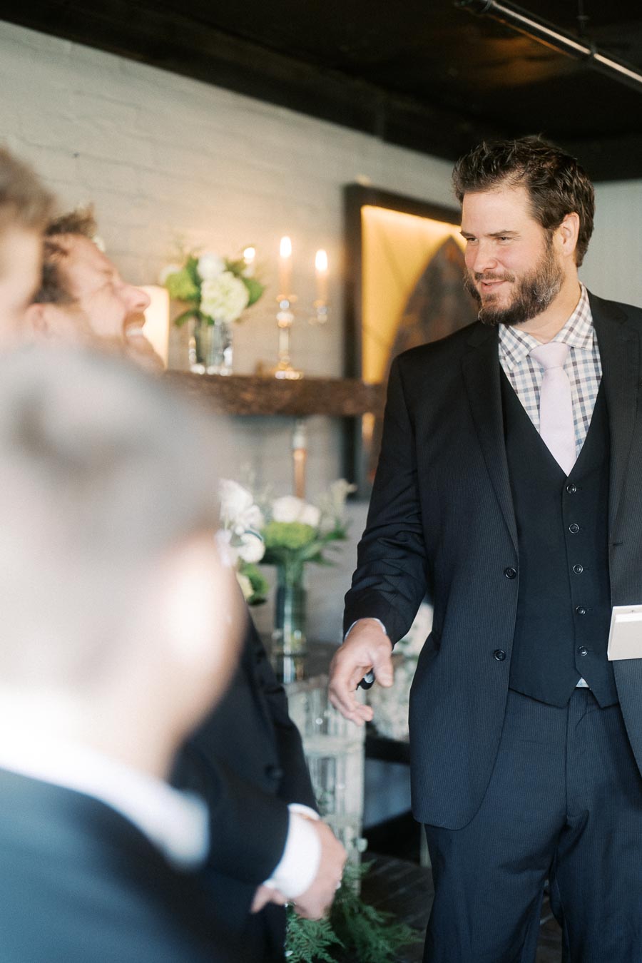 A man in a dark suit with a checkered shirt and light pink tie stands in a warmly lit room, engaging in conversation. The background features a rustic brick wall, decorative greenery, and lit candles, creating an elegant and cozy atmosphere.