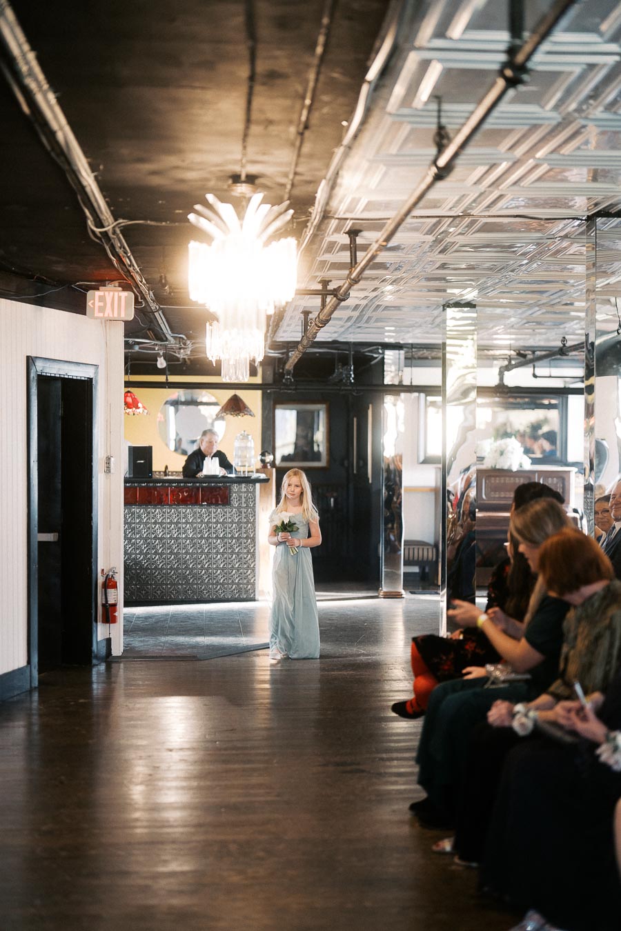 A young flower girl in a light blue dress walks down the aisle holding a bouquet during a wedding ceremony in an elegantly decorated venue with a mirrored ceiling and a chandelier, as guests watch.