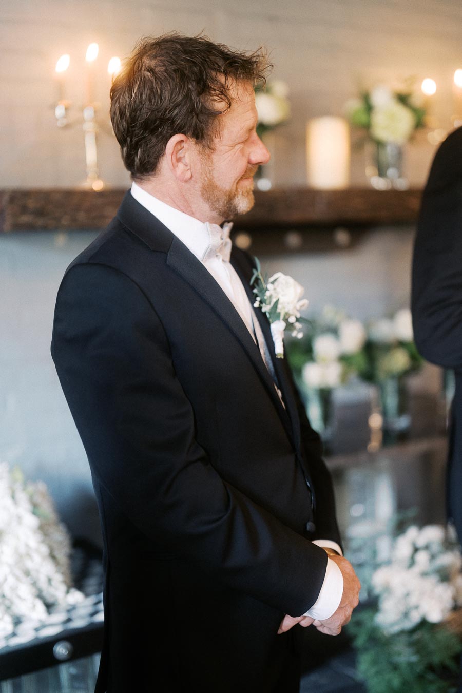Groom standing in formal black suit with white boutonniere, smiling at wedding ceremony in softly lit venue with floral decorations and candles.
