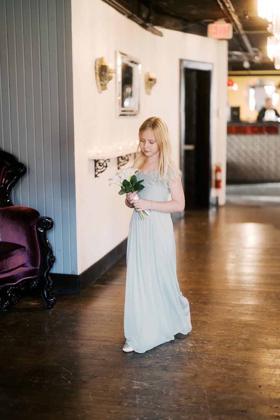A young girl in a light blue dress holding a bouquet of flowers walks gracefully in a warmly lit corridor with elegant decor.