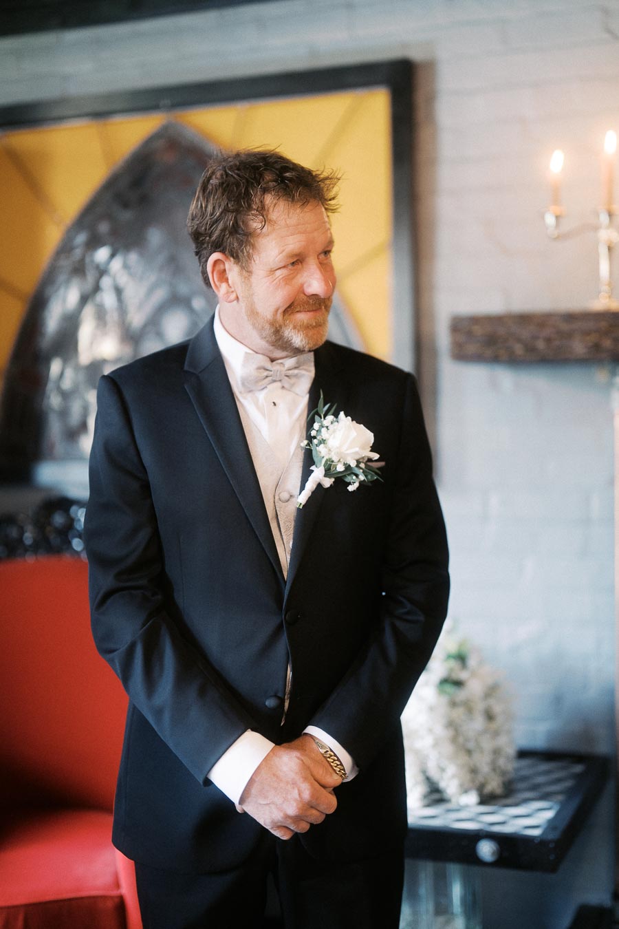 Man smiling in a formal black suit with a white boutonniere standing indoors next to a decorative backdrop and candlelight ambiance.
