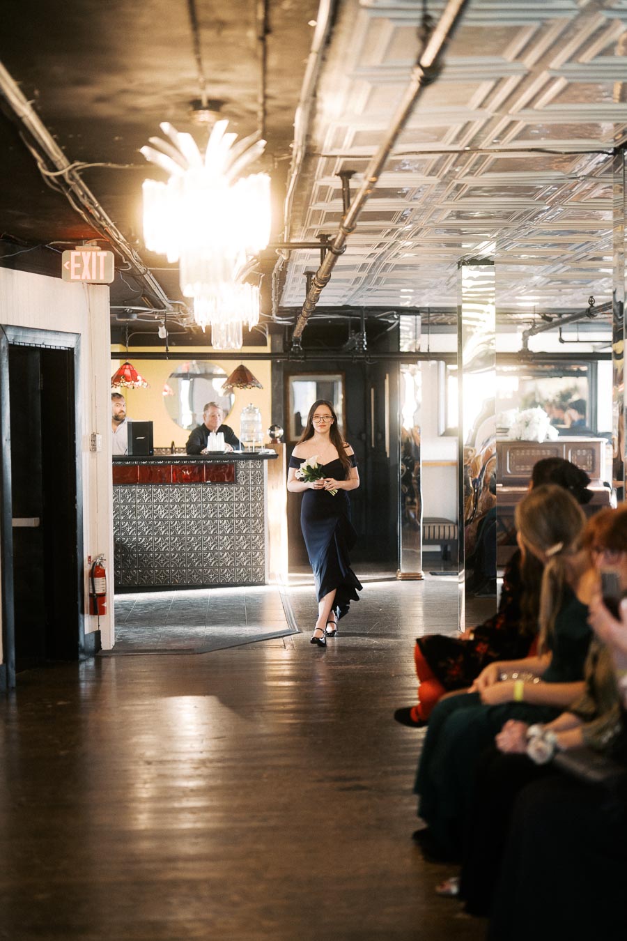 A woman in a navy blue dress walks down an indoor aisle holding a bouquet, with an elegant chandelier overhead and guests seated to her right.