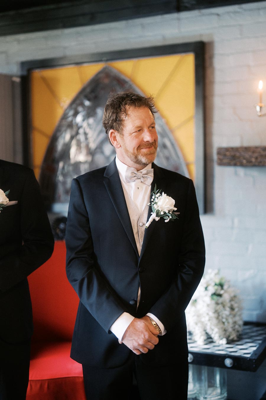 A middle-aged man in a black suit with a bow tie and boutonniere, smiling during a formal event, stands indoors by a stained glass window and white brick wall, with a red chair and floral decorations in the background.