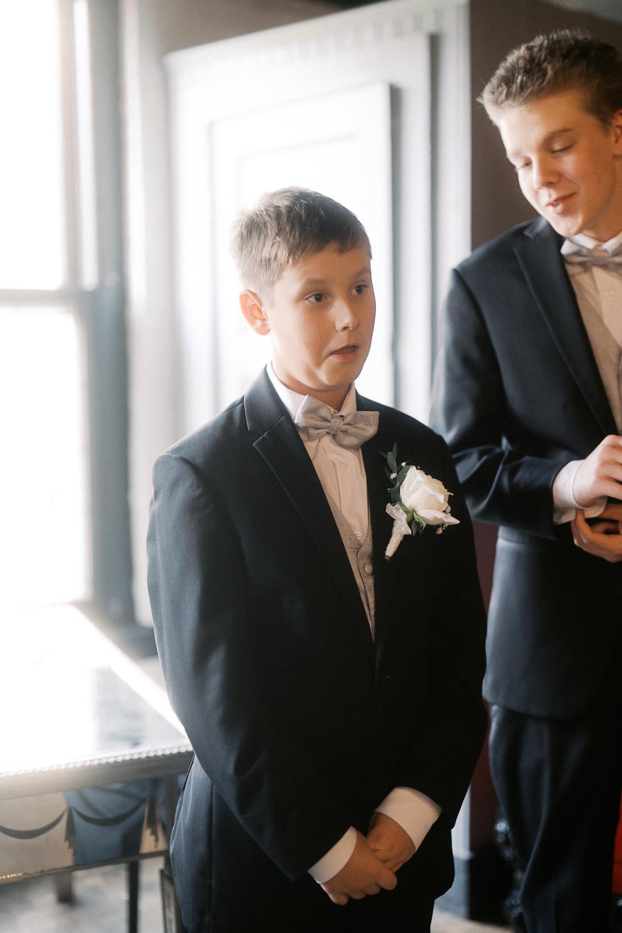 A young boy in a formal black suit and bow tie, with a white floral boutonniere, standing indoors next to another person in similar attire, possibly at a wedding or formal event.