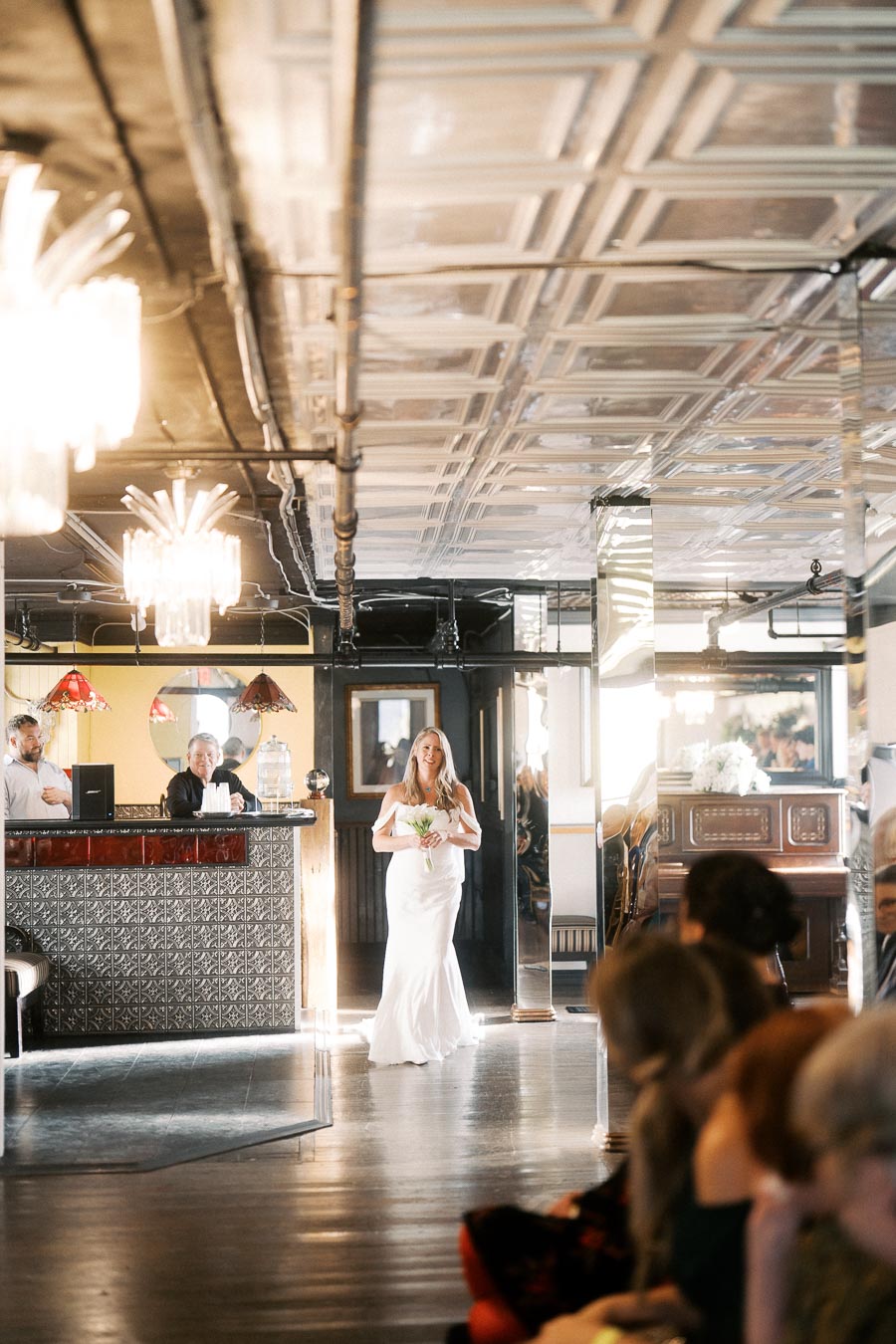 Elegant bride in white dress walking down the aisle in a stylish, warmly lit venue, holding a bouquet; guests seated, awaiting her arrival.