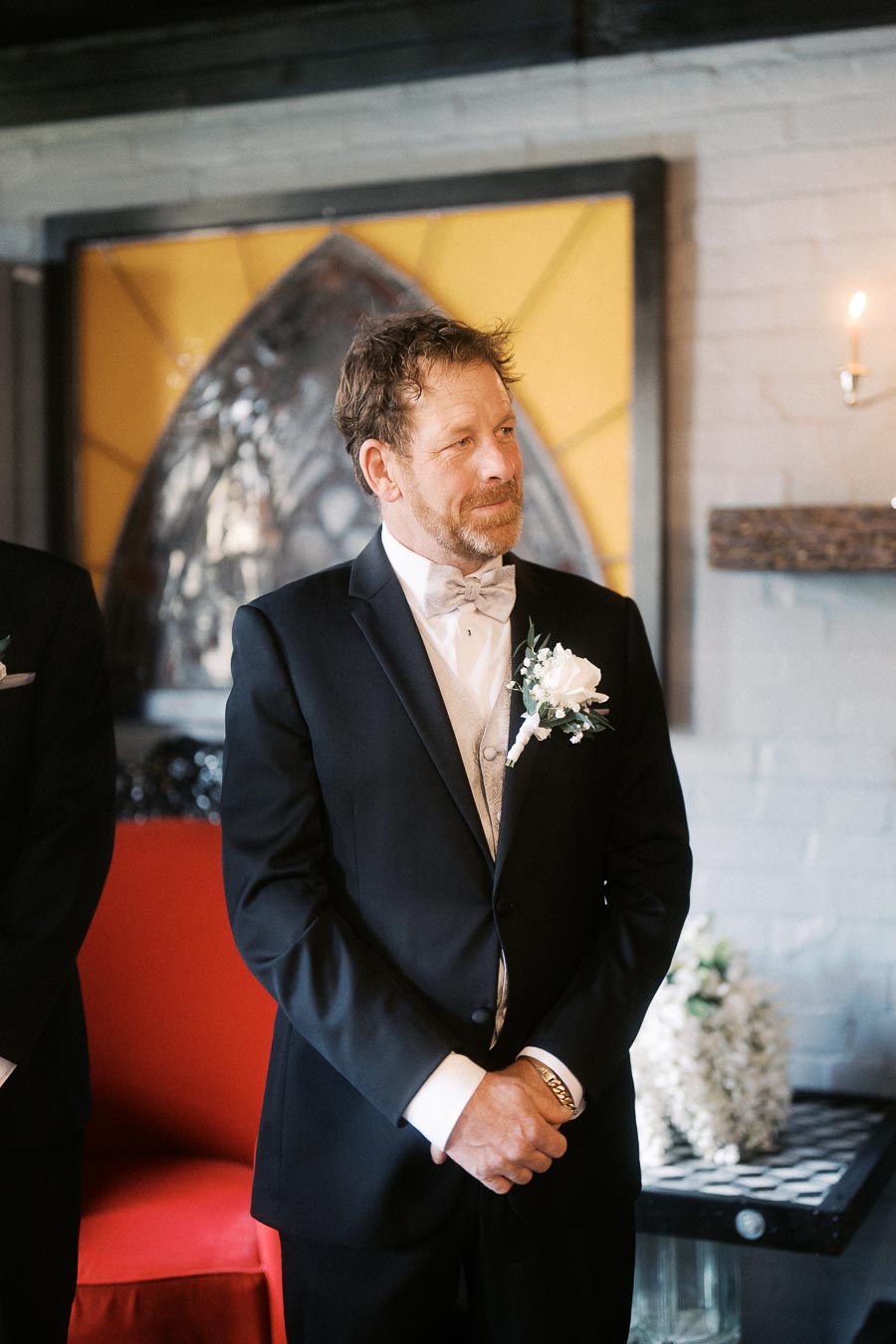 A groom in a formal tuxedo stands indoors at a wedding ceremony, with a boutonniere and a gentle smile, framed by elegant decor.