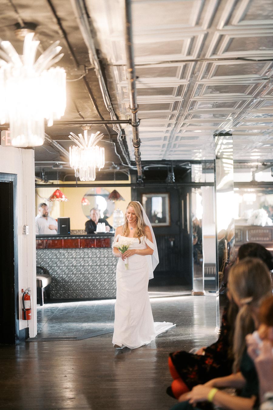 A bride in a white dress holding a bouquet walks down a dimly lit aisle in a stylish venue with chandeliers and guests seated on the side.