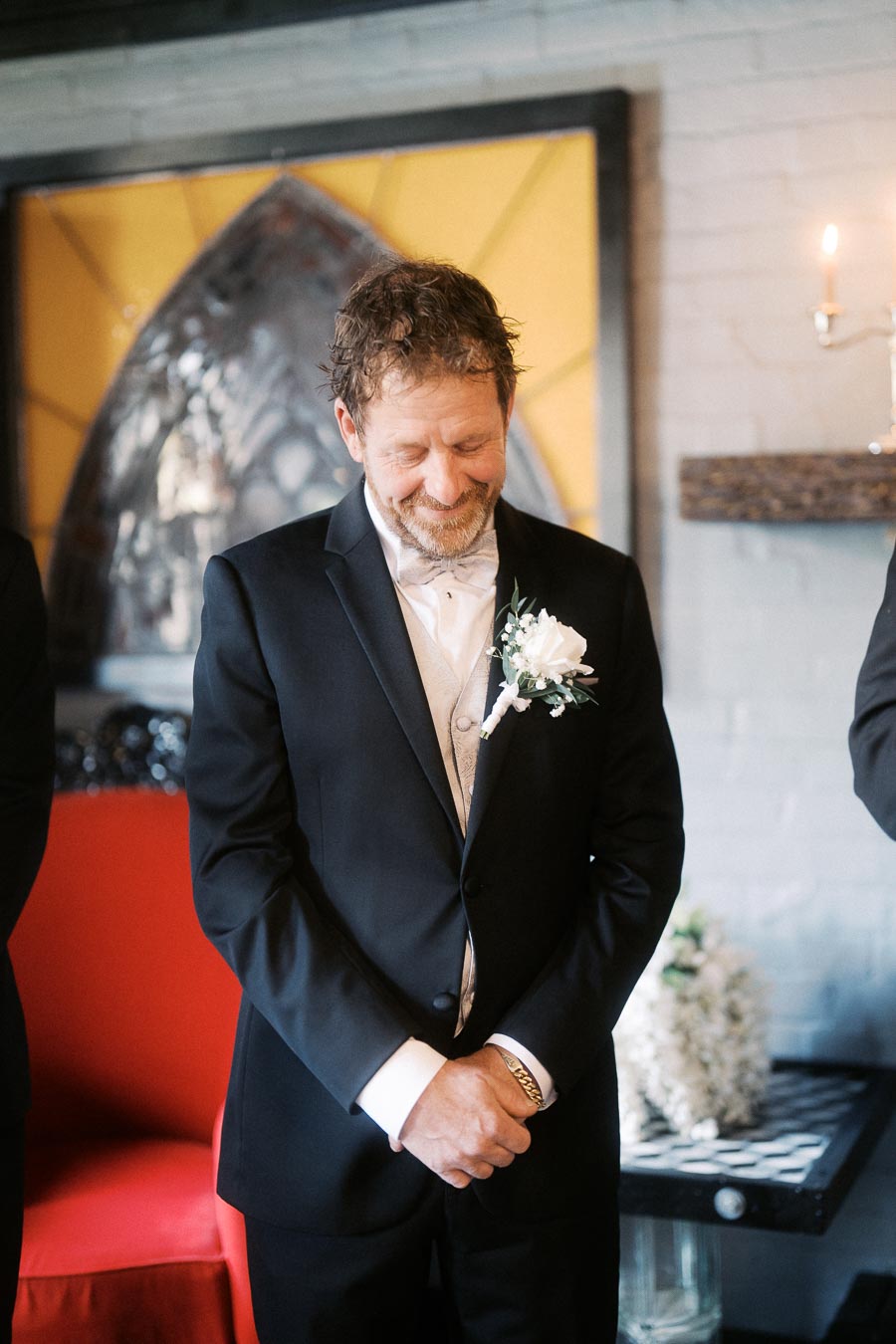 Middle-aged man in a black suit with a white boutonniere, standing with eyes closed and smiling, in a formal indoor setting with decorative background elements.