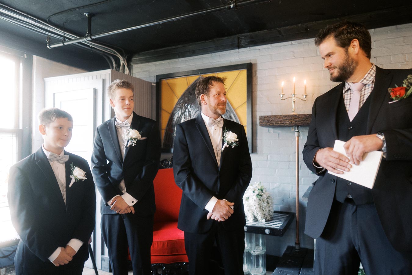 Groom and groomsmen standing in a stylish, dimly lit room with formal attire, adorned with white boutonnieres, during a wedding ceremony.