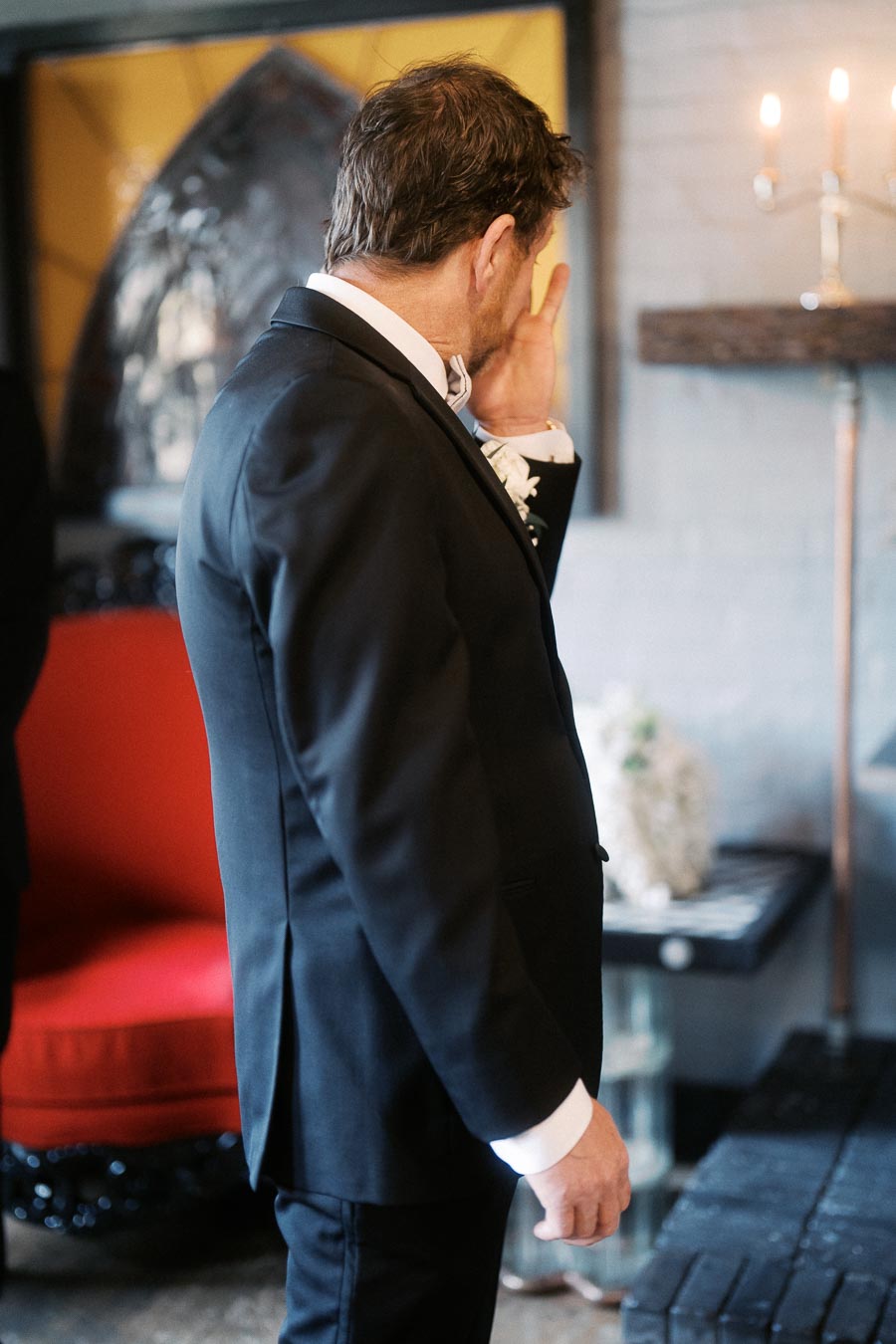 A man in a suit wiping a tear from his eye, standing in an elegantly decorated room with a red chair and a lit candelabra in the background, evoking a poignant emotional moment.