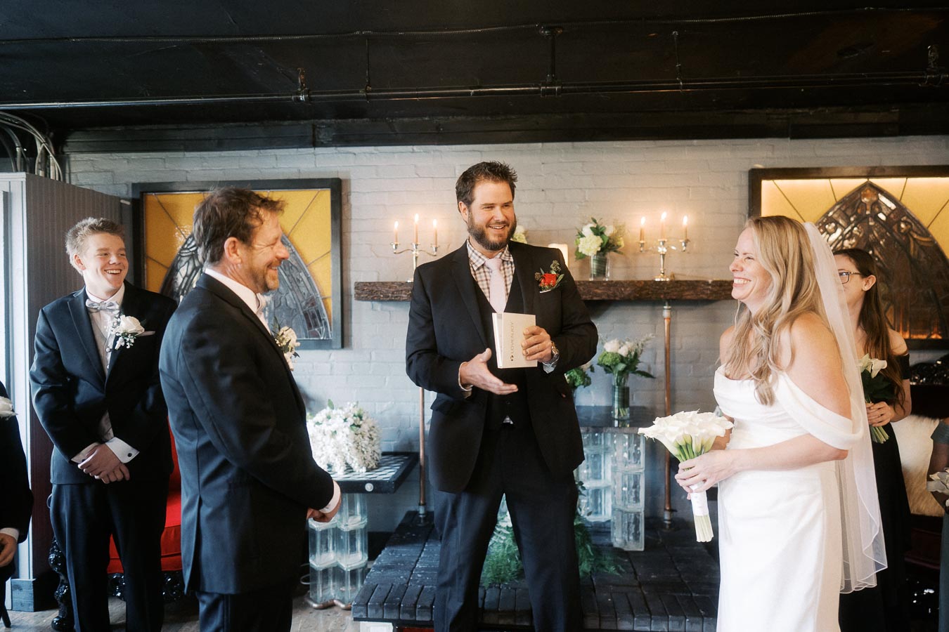 Wedding ceremony in a cozy venue with a smiling bride in a white dress holding a bouquet, surrounded by joyful guests and a speaker in a suit holding an open book, with elegant decor and lit candles in the background.