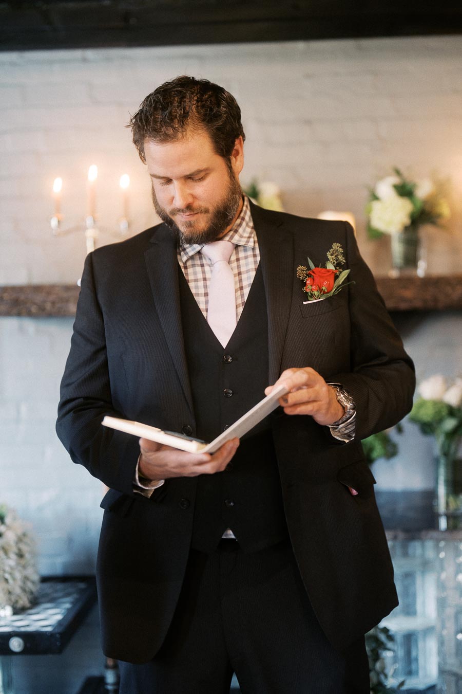 Man in a suit with a pink tie reading from a book in an elegant setting with flowers and candles.