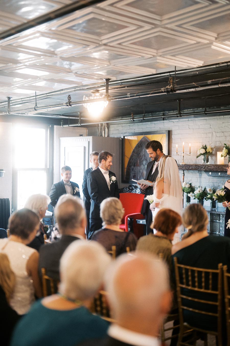 Wedding ceremony in a vintage venue with groom and officiant standing, guests seated in foreground, elegant decor with floral arrangements and candles in the background.
