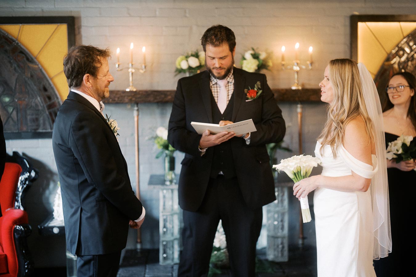 Couple exchanging vows in an elegant indoor wedding ceremony with officiant, surrounded by floral decorations and candlelight.