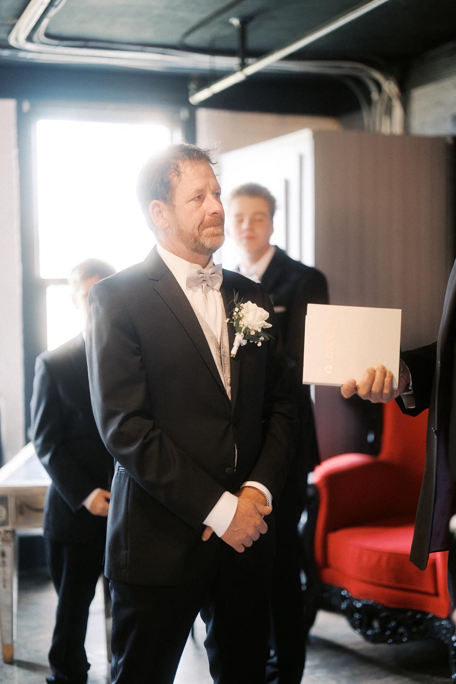 A man in a suit stands solemnly next to another man holding a document, with a red chair in the background, suggesting a formal occasion or event.