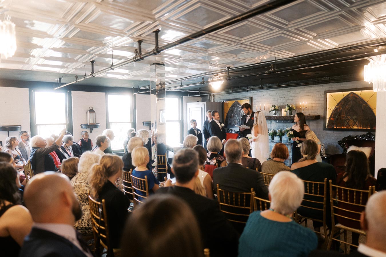 Indoor wedding ceremony with guests seated on golden chairs, facing a couple exchanging vows in a warmly decorated venue with chandeliers and large windows.