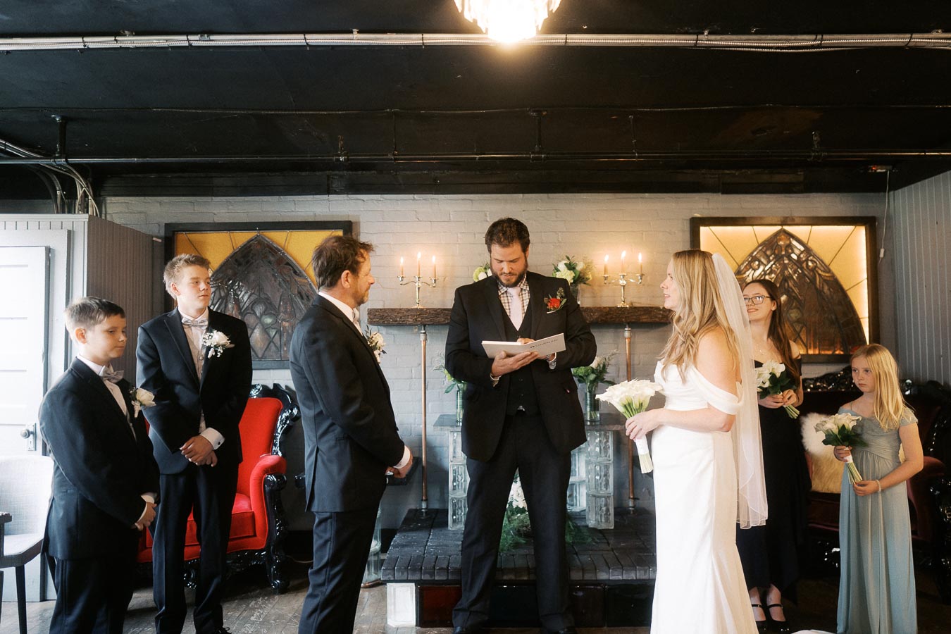 A small wedding ceremony takes place indoors with a bride in a white gown and veil, a groom, and young groomsmen in black suits. A man officiates in front of a decorated mantle with lit candles, while bridesmaids in dark dresses, one holding flowers, stand nearby. The setting is intimate and elegantly decorated.