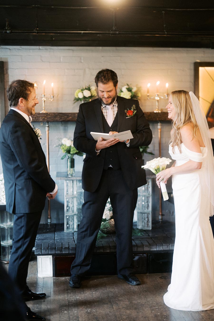Bride and groom exchanging vows during an intimate indoor wedding ceremony, with the officiant smiling and candlelit ambiance in the background.