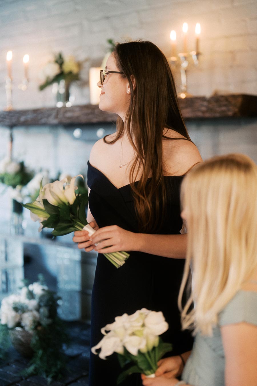 Elegant woman in black dress holding a bouquet of white calla lilies, standing beside a young girl in a light dress, with a softly lit, decorated mantel in the background.
