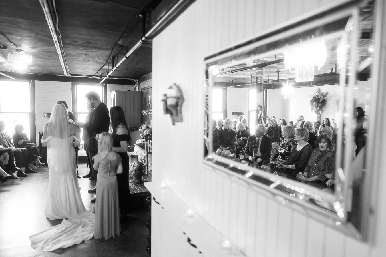 Black and white image of an intimate indoor wedding ceremony. The bride and groom stand together at the altar, accompanied by two children in elegant dresses. Guests are seated attentively, captured in a large mirror reflection. The venue features large windows and soft, ambient lighting.