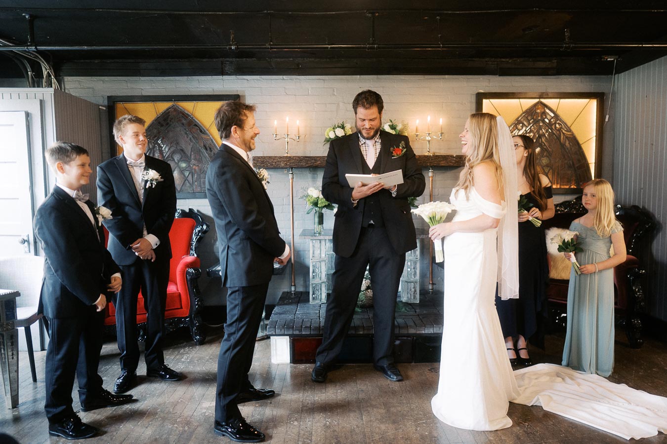 Indoor wedding ceremony with a bride and groom, surrounded by attendants, standing before an officiant. The room is decorated with flower arrangements and lit candles, creating an intimate and elegant atmosphere.