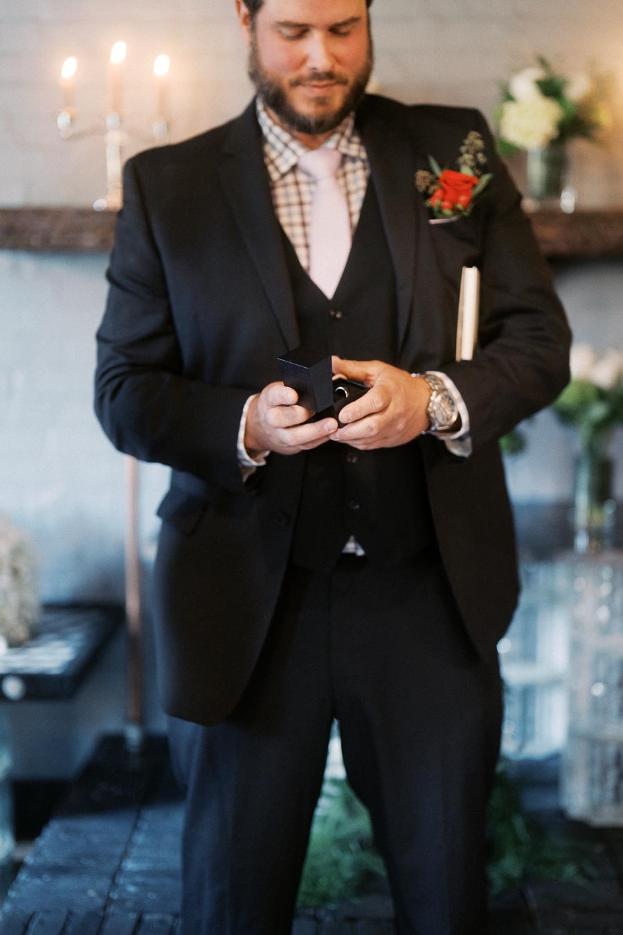 Man in a suit holding a small box, possibly for a proposal, with flowers and candles in the background at an indoor event.