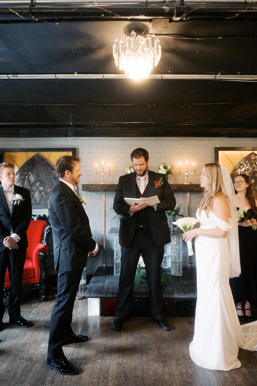 Elegant indoor wedding ceremony with groom, bride in white gown holding bouquet, and officiant reading vows under chandelier lighting.