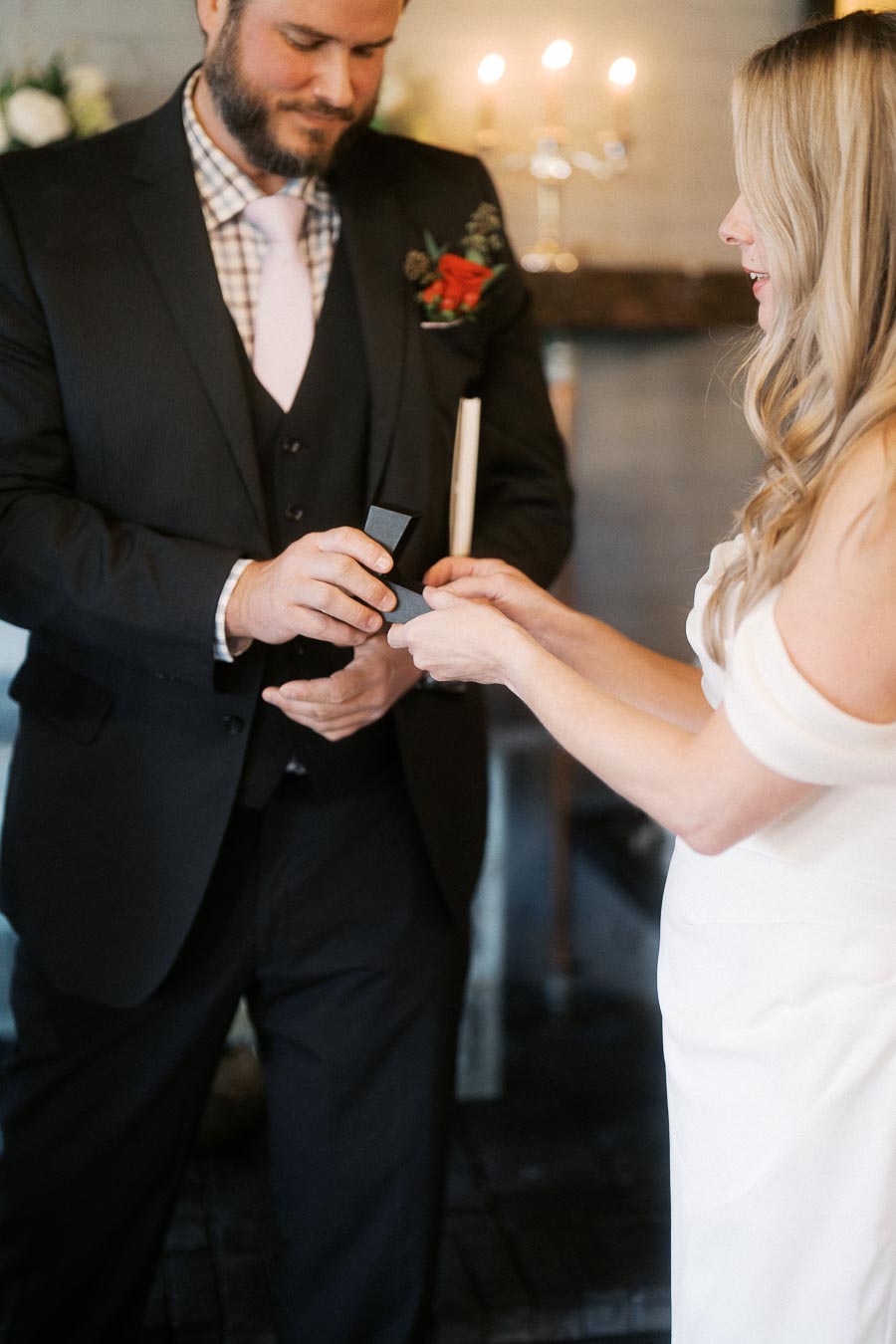 A couple exchanging wedding rings during a ceremony, with the groom in a black suit and the bride in a white dress, symbolizing love and commitment.