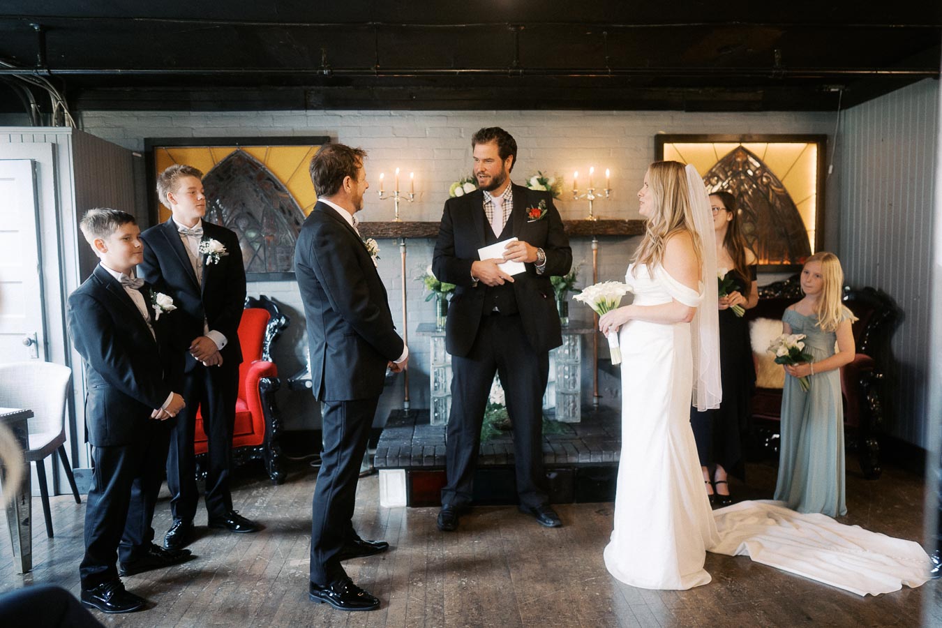 Elegant indoor wedding ceremony with a bride in a white dress holding flowers and a groom in a black suit. An officiant stands between them reading vows. Bridesmaids and groomsmen in formal attire observe. Romantic ambiance with candles and decor in the background.