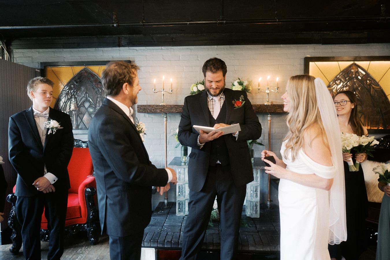 Wedding ceremony with a smiling bride in a white dress and veil, facing the groom in a black suit. The officiant is reading from a book, with bridesmaids holding bouquets and groomsmen attentively observing in a warmly lit venue.