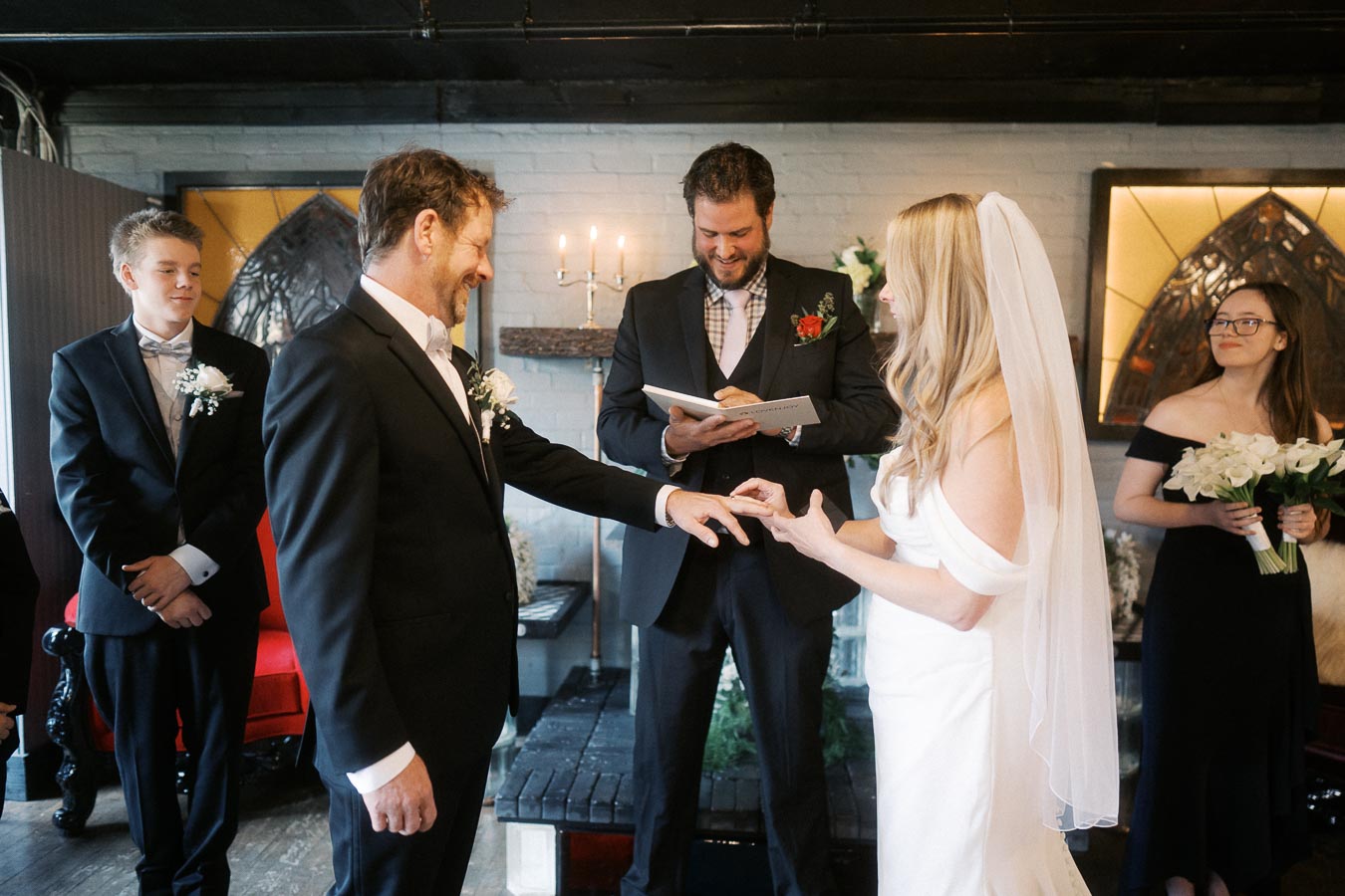 Bride and groom exchanging rings during a wedding ceremony, attended by a celebrant and two witnesses.
