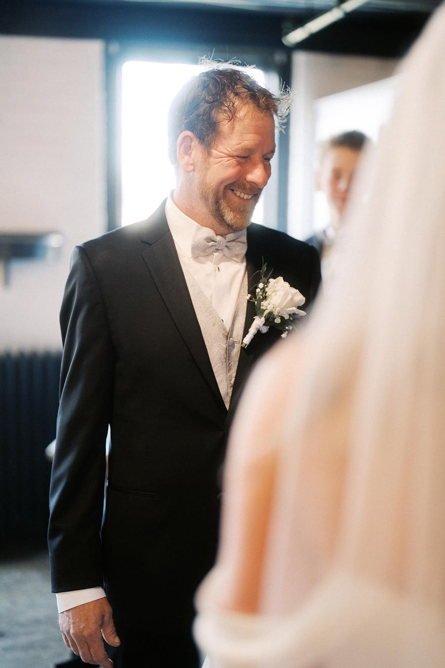 A groom smiling warmly at his bride during a wedding ceremony, wearing a black suit with a white shirt and bow tie, complemented by a floral boutonniere.