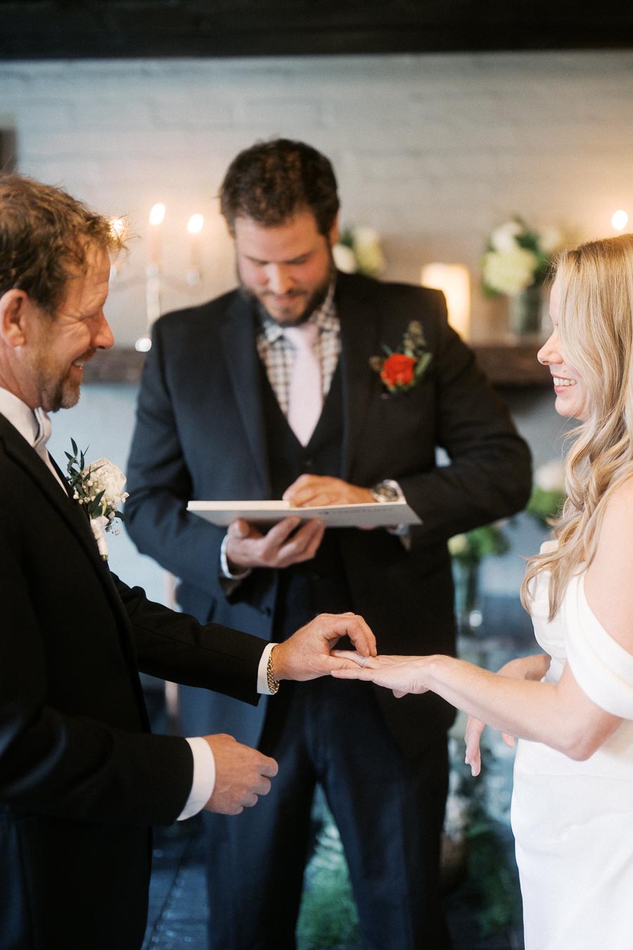 Wedding ceremony with groom placing ring on bride's finger, officiant holding vows in background, romantic setting with candles and flowers.